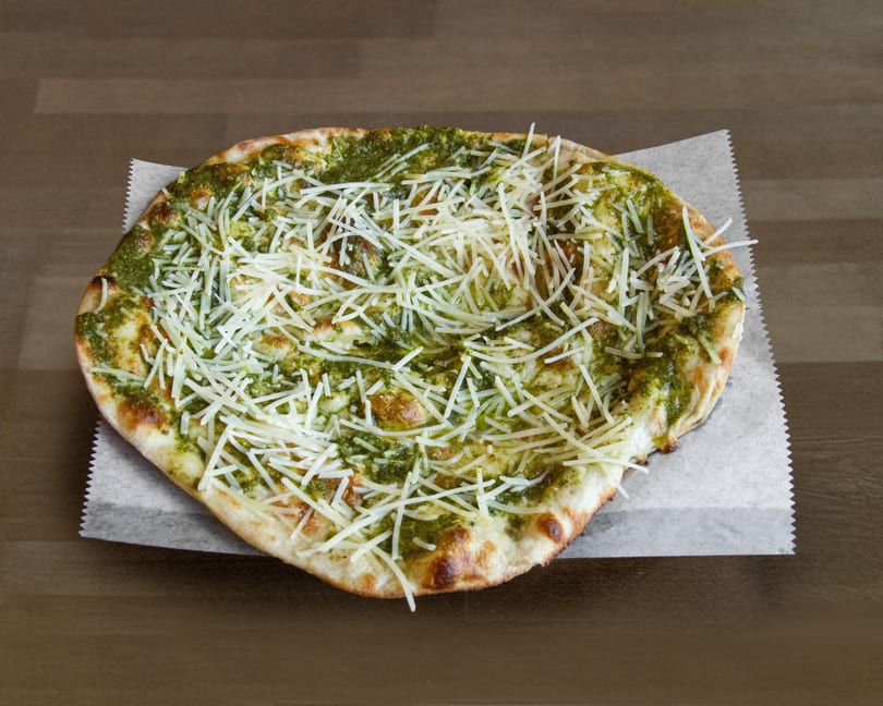 A round pesto flatbread pizza, topped with shredded white cheese, on a piece of parchment paper.