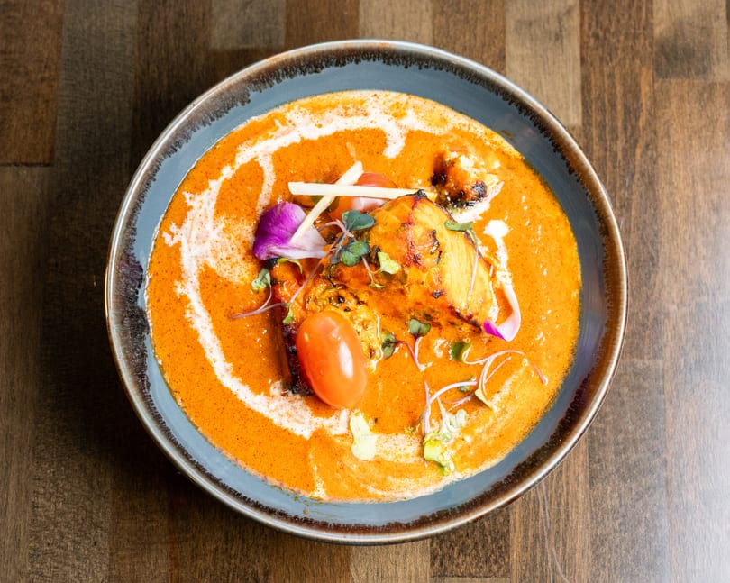 Overhead view of a vibrant orange curry dish with grilled chicken, cherry tomato, and garnishes.