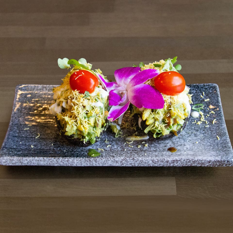 Two stuffed avocado halves topped with cherry tomatoes, microgreens, and a purple orchid on a grey plate.