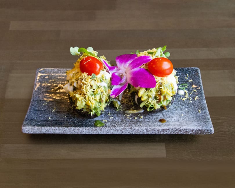 Two stuffed avocado halves topped with cherry tomatoes, microgreens, and a purple orchid on a grey plate.