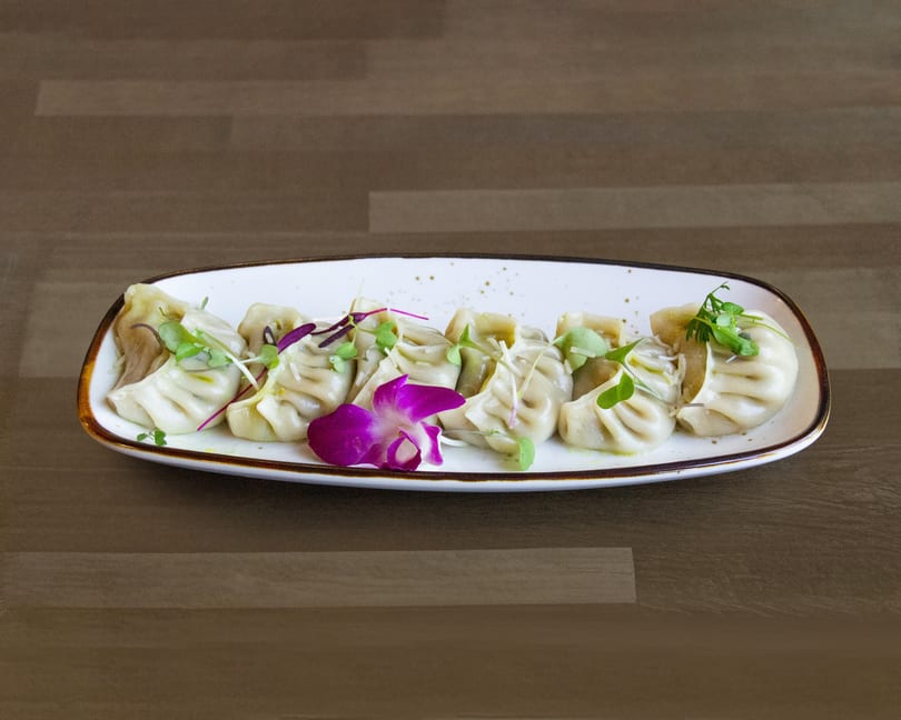 A white rectangular plate of dumplings garnished with microgreens and a purple orchid.