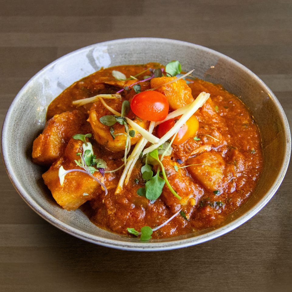 A bowl of Indian curry with paneer, cherry tomatoes, and microgreens on a wooden table.