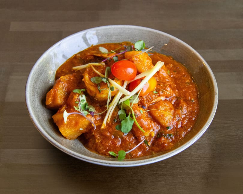 A bowl of Indian curry with paneer, cherry tomatoes, and microgreens on a wooden table.