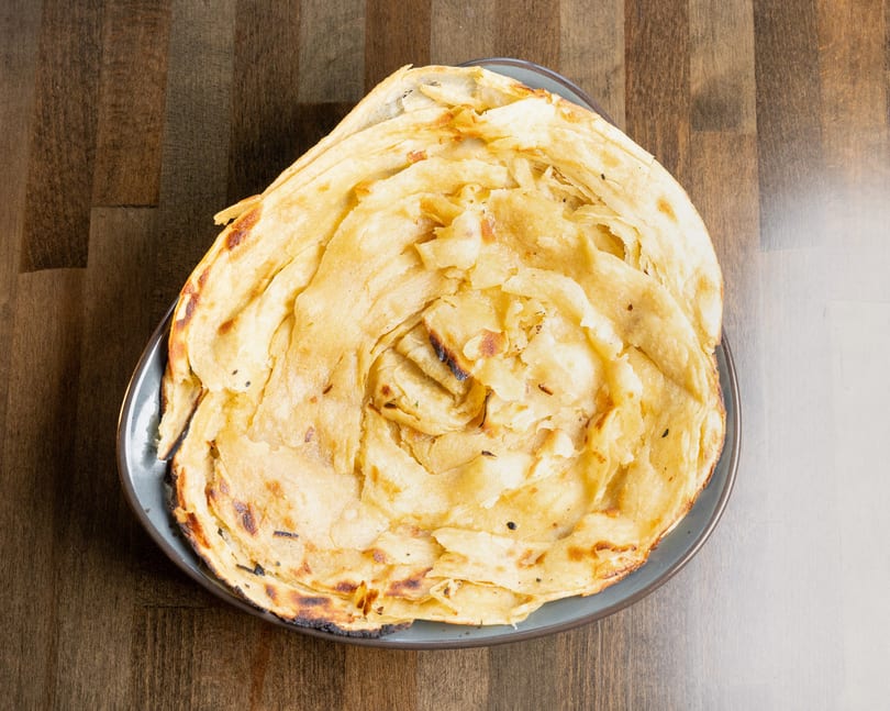 A plate of golden-brown, flaky paratha bread, lightly charred, on a wooden table.