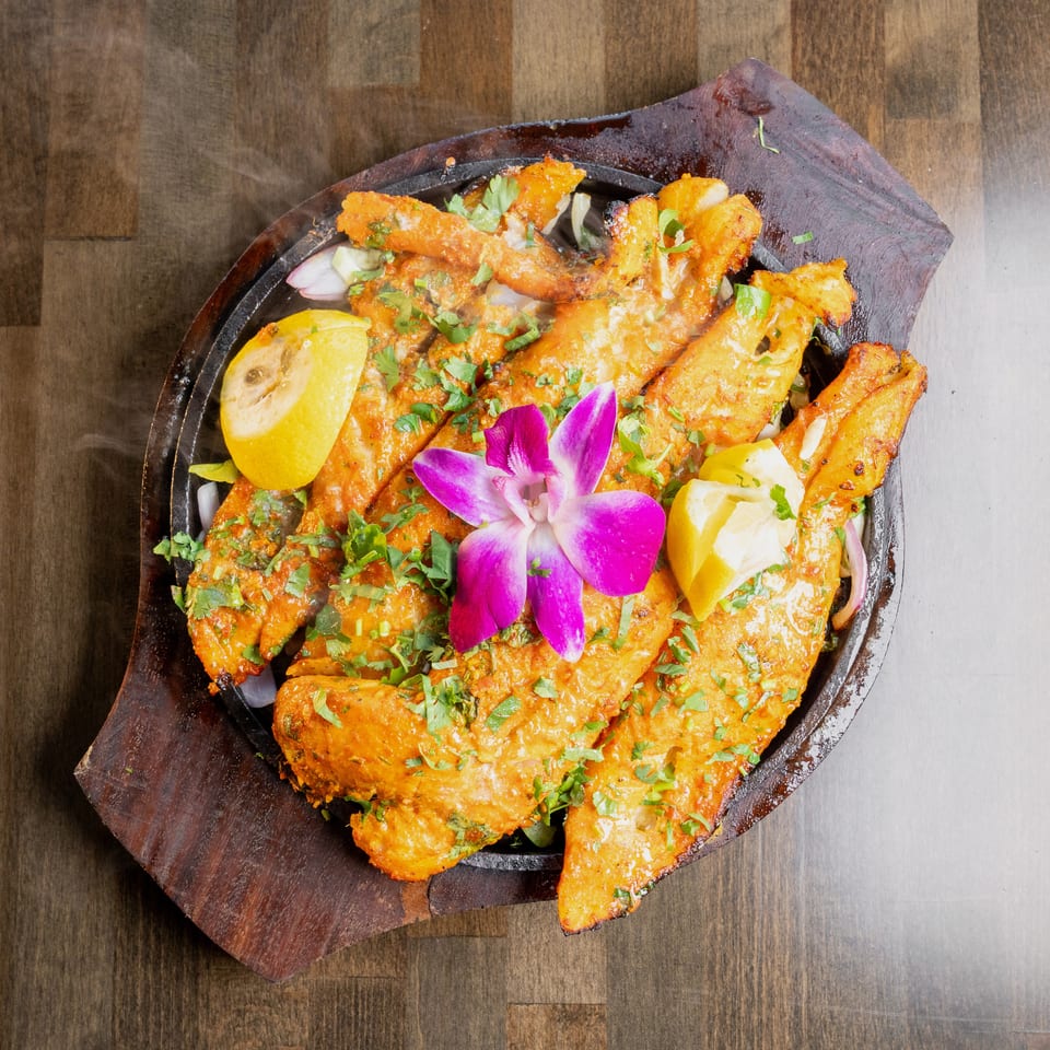 Sizzling orange fish fillets garnished with cilantro, lemon, and a purple orchid on a wooden platter.