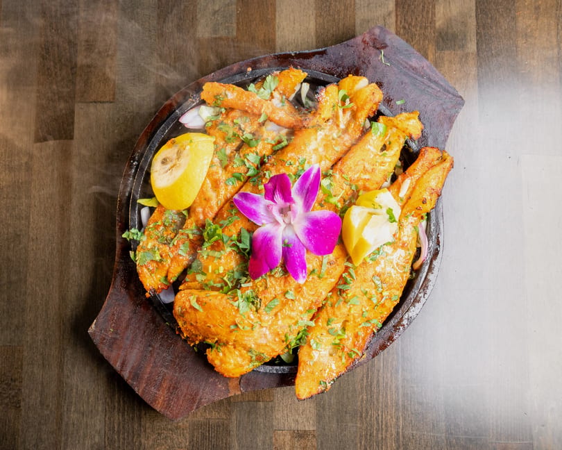 Sizzling orange fish fillets garnished with cilantro, lemon, and a purple orchid on a wooden platter.