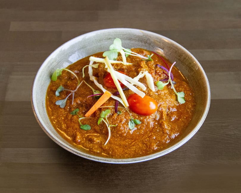 A bowl of rich, orange-brown curry garnished with microgreens, cherry tomatoes, and julienned vegetables.