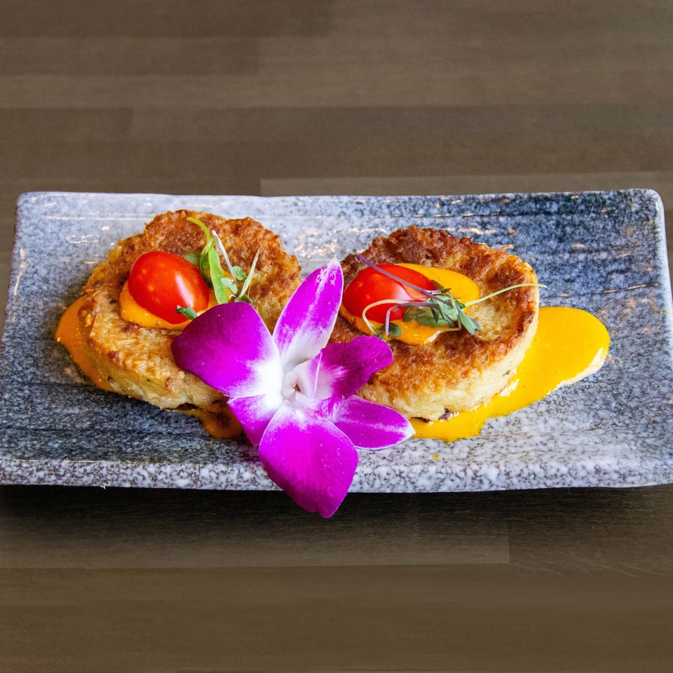 Two golden-brown crab cakes with cherry tomatoes, microgreens, and yellow sauce, garnished with a purple orchid.
