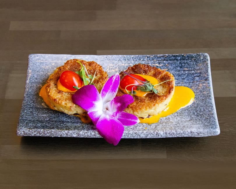 Two golden-brown crab cakes with cherry tomatoes, microgreens, and yellow sauce, garnished with a purple orchid.