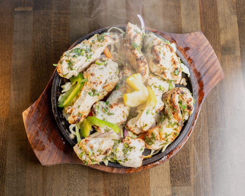 Sizzling chicken tikka with vegetables and lemon wedges on a cast iron platter.