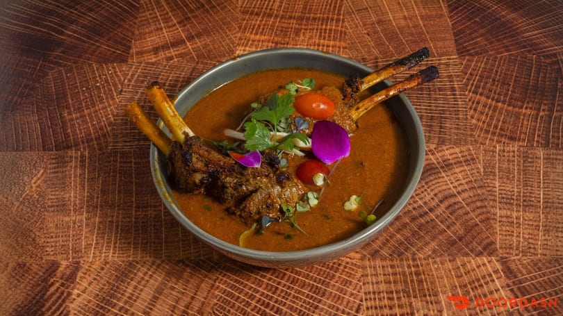 Lamb shank curry with cherry tomatoes, cilantro, and edible flowers in a gray bowl.