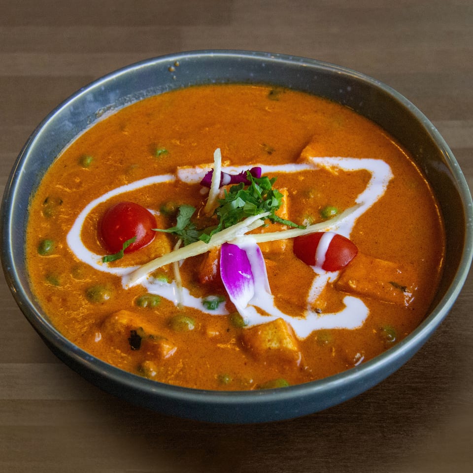 A vibrant orange curry with peas, paneer, cherry tomatoes, and a swirl of cream in a dark bowl.