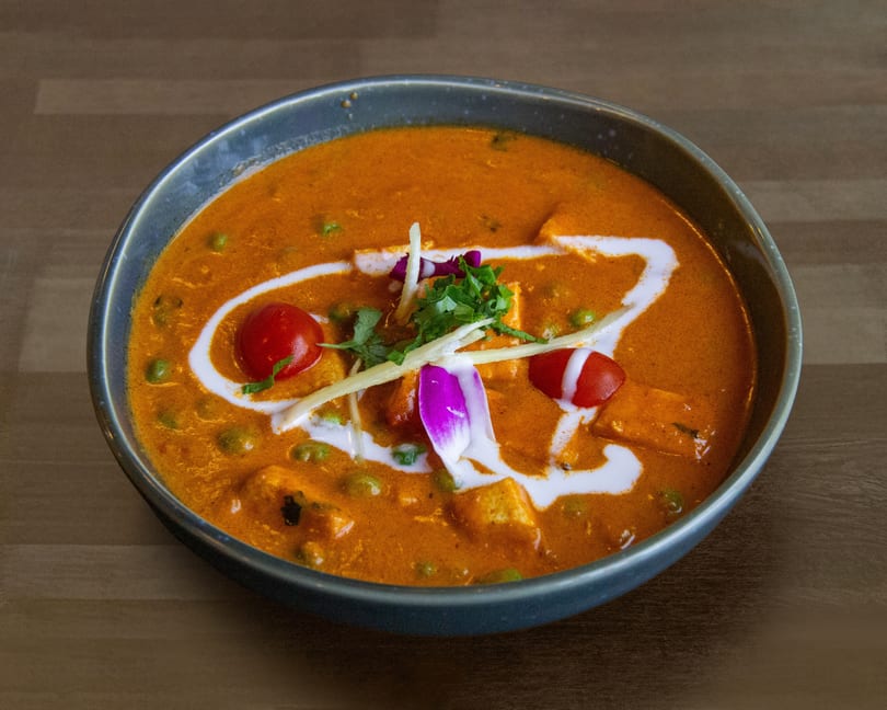 A vibrant orange curry with peas, paneer, cherry tomatoes, and a swirl of cream in a dark bowl.
