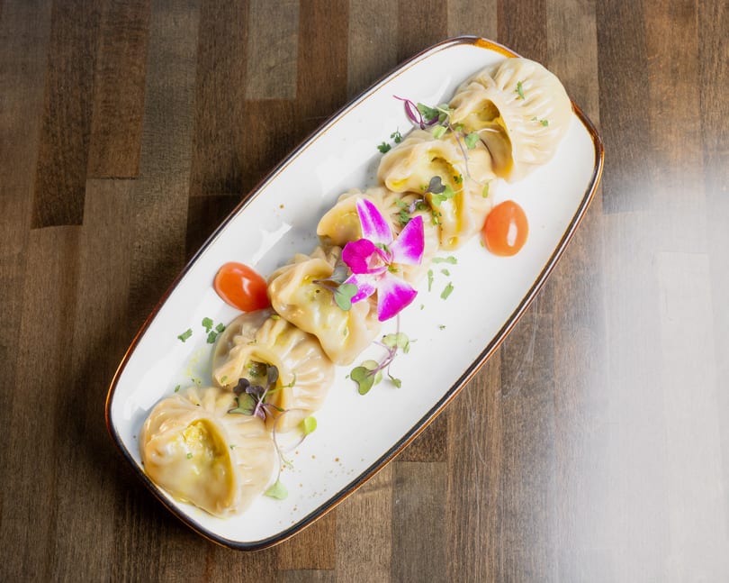 Steamed dumplings, garnished with microgreens, cherry tomatoes, and an orchid, on a white plate.