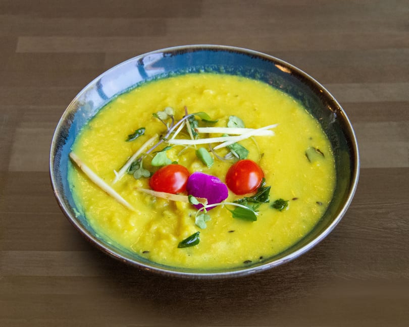A vibrant yellow lentil dal in a blue bowl, garnished with cherry tomatoes, herbs, and ginger.