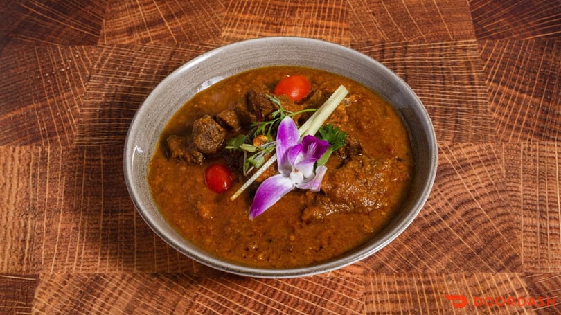 Rich, orange curry with meat, cherry tomatoes, and a purple orchid in a gray bowl on wood.