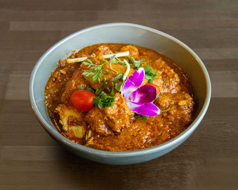 Chicken curry in a bowl, garnished with cilantro, cherry tomatoes, and a purple orchid.