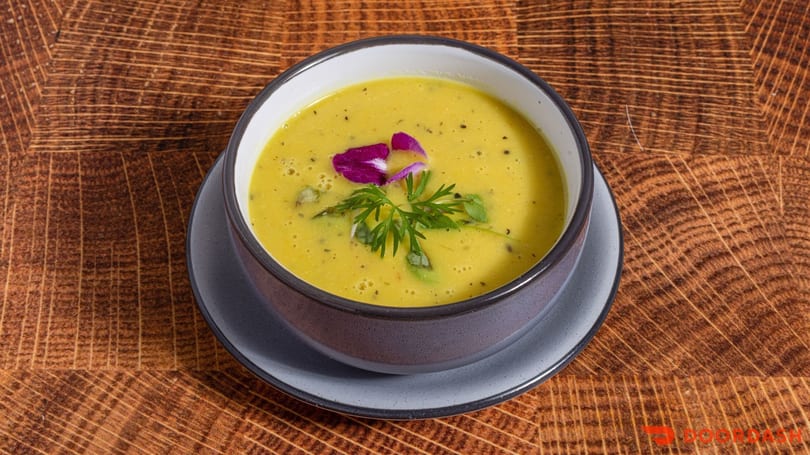 Yellow soup in a dark bowl, garnished with a purple flower and green herbs on a wooden table.
