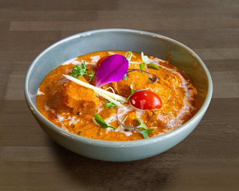 A bowl of orange curry with cream swirls, garnished with microgreens, cherry tomatoes, and a purple orchid petal.