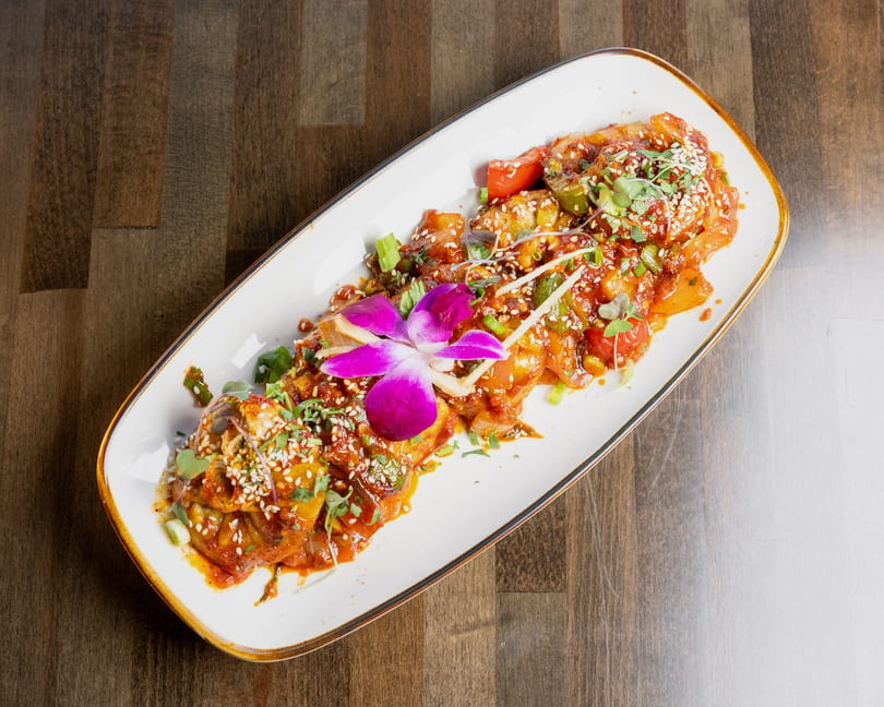 A long white plate of chili paneer, garnished with sesame seeds, cilantro, and an orchid.