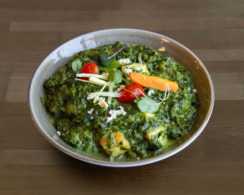 A bowl of vibrant green palak paneer, garnished with tomatoes, carrots, and fresh herbs.